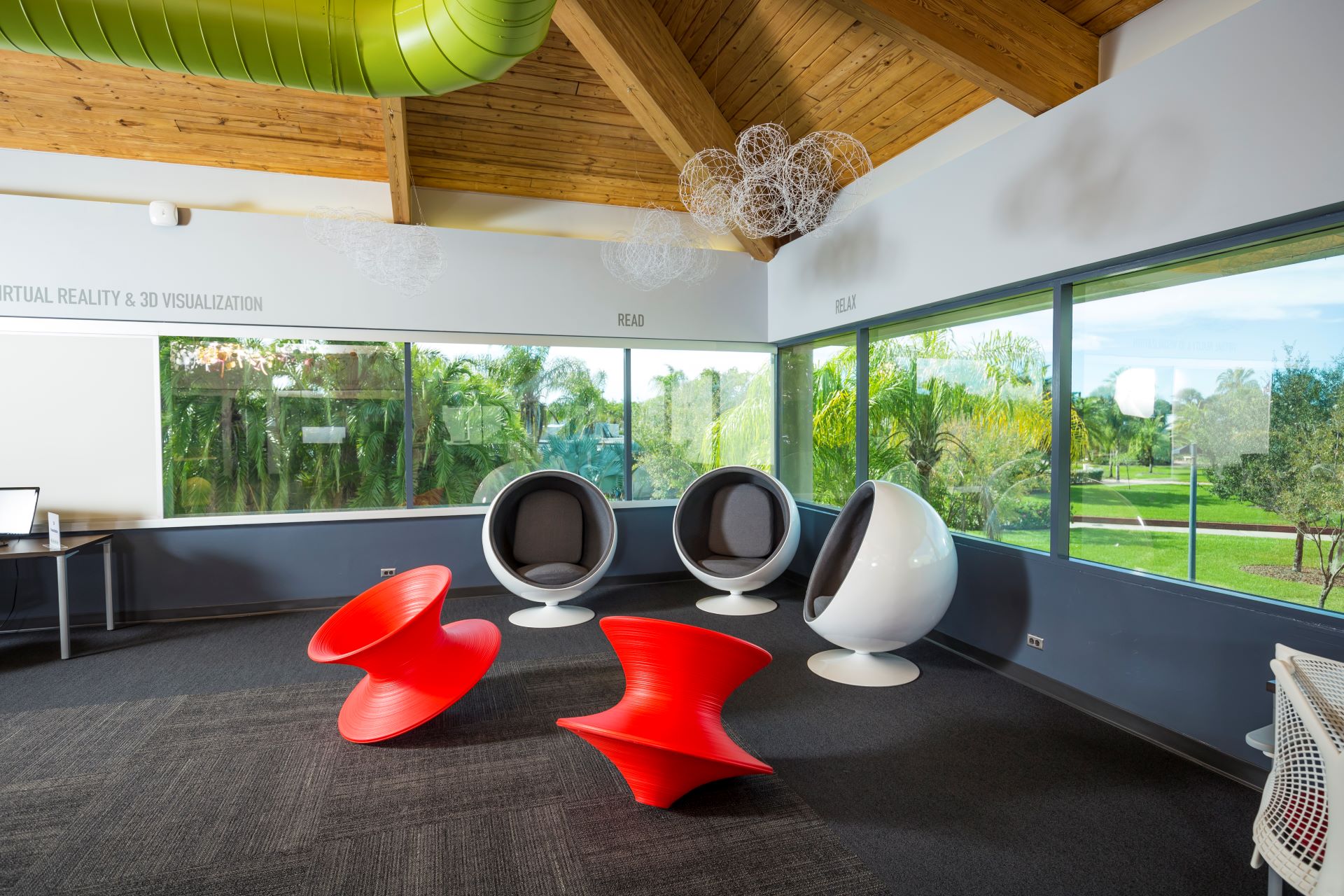 Interior view of a modern university lounge designed for relaxation and study, featuring a high wooden ceiling with green beams and intricate white light fixtures. The area includes unique red and white seating options, large windows showcasing a lush garden view, and labeled zones for reading and relaxing